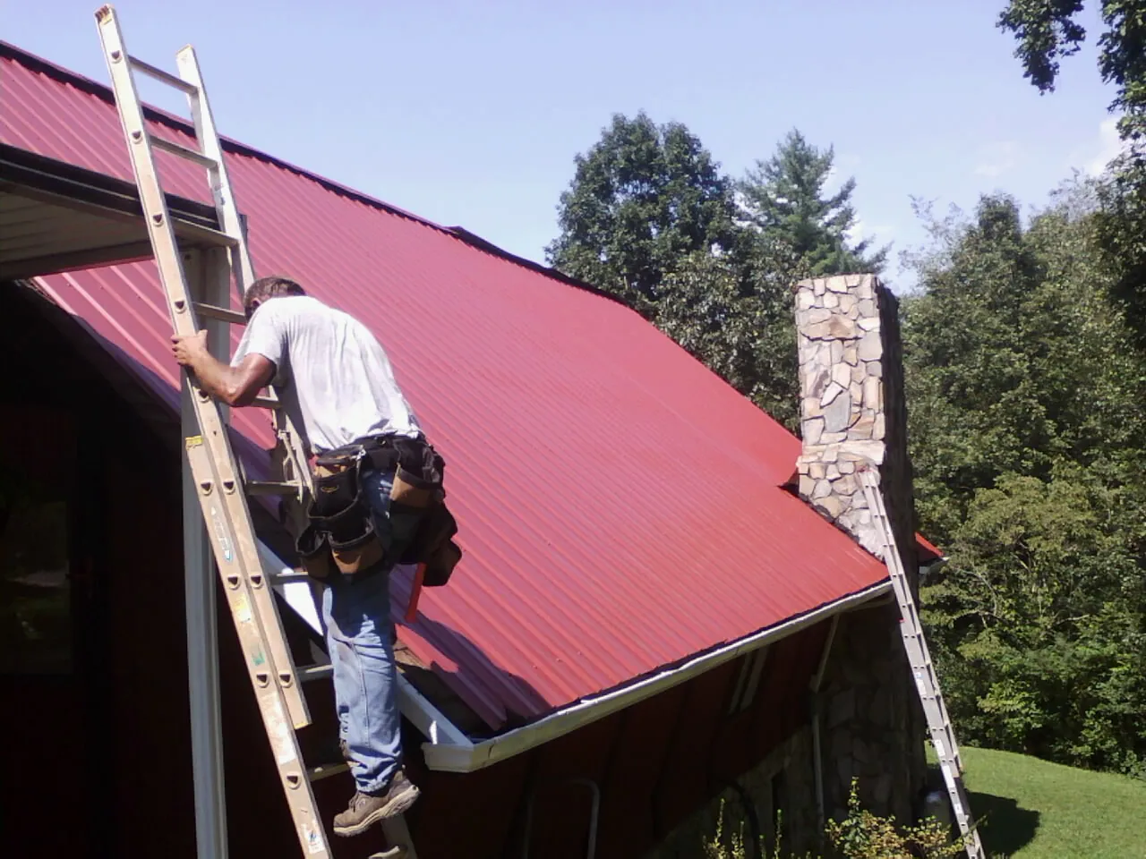 Roofer on a ladder beside a red metal roof and stone chimney — Soffit & Fascia Repair by Roofing Brothers in Miramar Beach